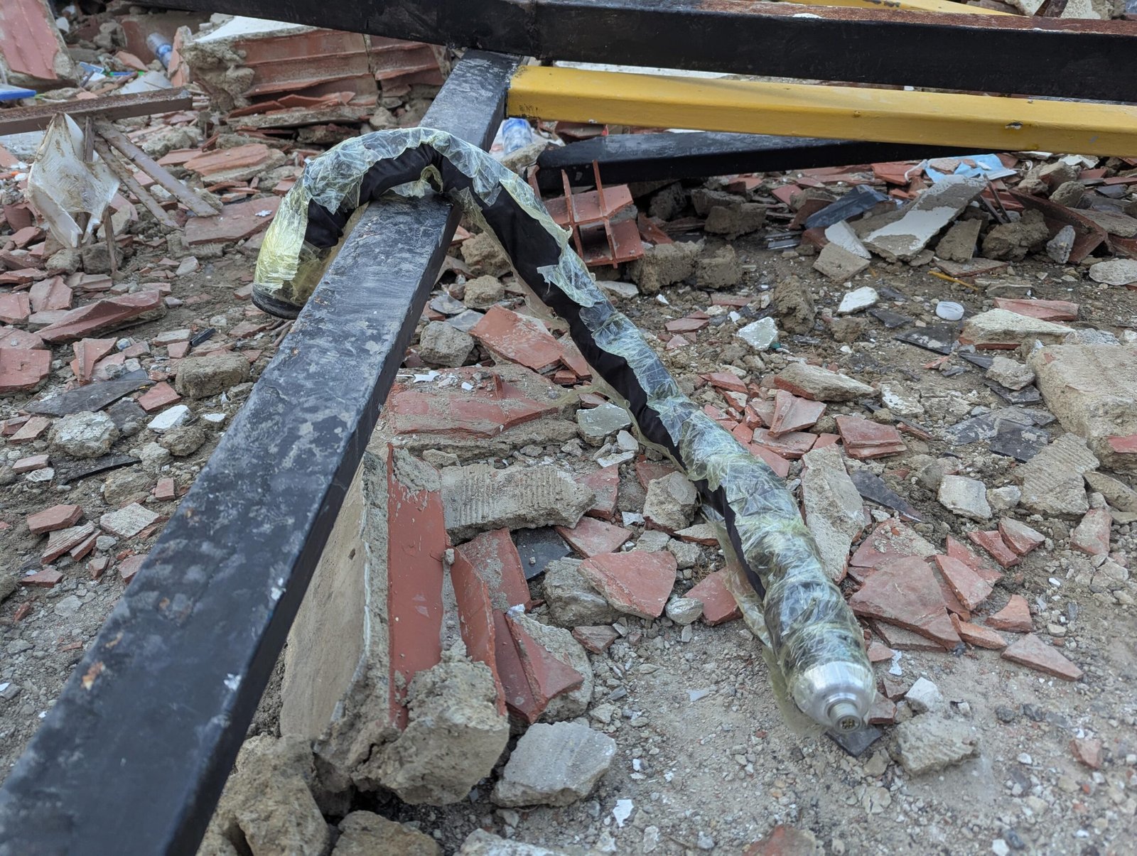 A long black snake robot covered in plastic material lies on top of a bar amid building debris from an earthquake.