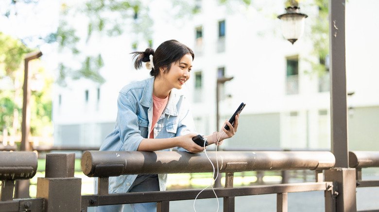 A woman leaning against a railing and enjoying a video call on a phone while it's plugged into a charger.
