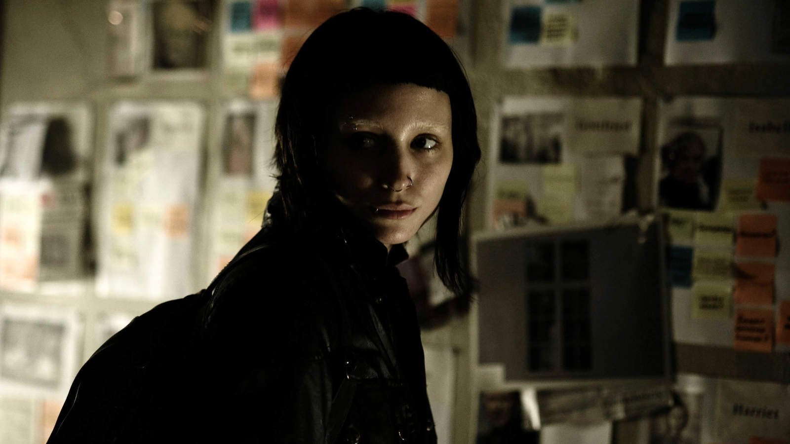 Rooney Mara's Lisbeth looking over her shoulder in front of a wall with papers taped to it in The Girl with the Dragon Tattoo