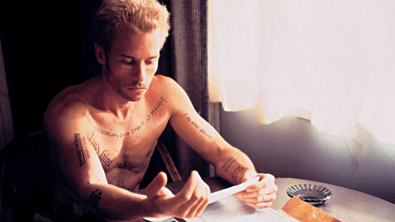Leonard Shelby (Guy Peace) a shirtless young man with blonde hair covered in text tattoos, sits at a table in a hotel room and reads a note