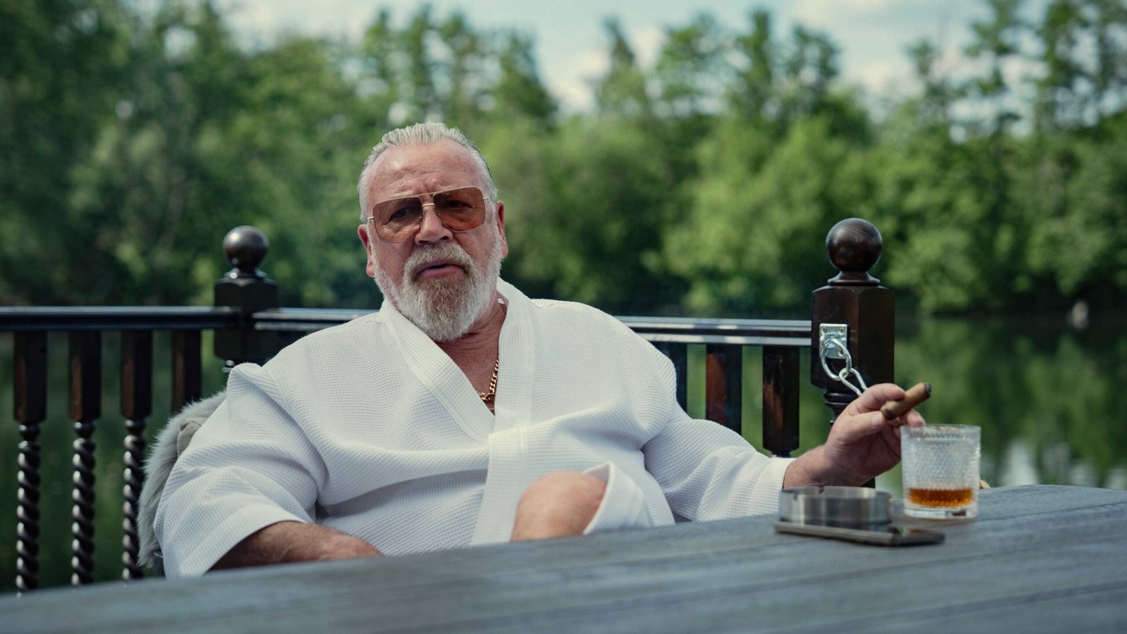 Bobby Glass (Ray Winstone) sits with a drink outside with a lake behind him in The Gentlemen season 2