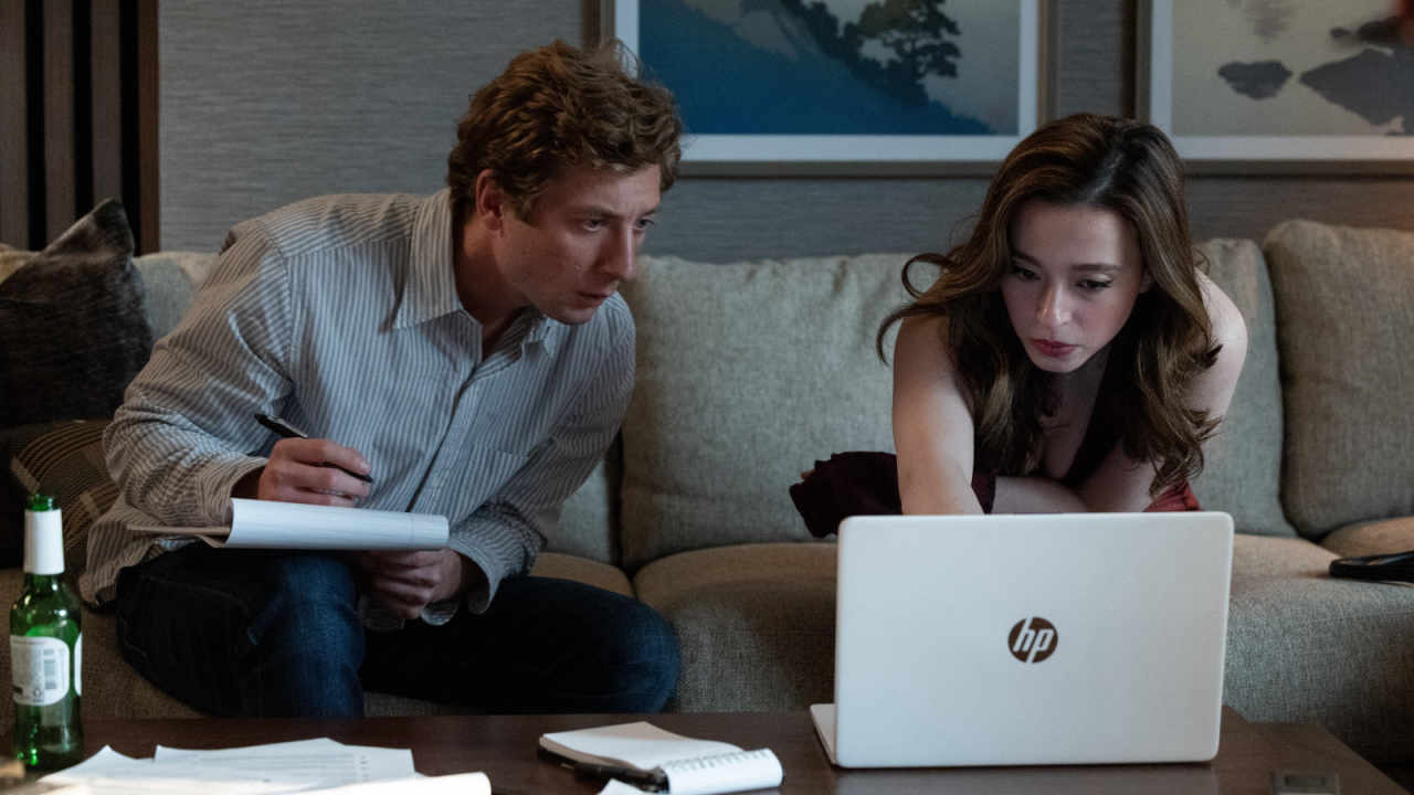 A man in a pinstripe button down shirt sitting on a couch with a woman in a cocktail dress. The pair are leaning forward to look at a laptop that’s placed in front of them on a table.